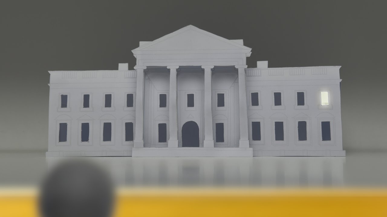 Das Weisse Haus aus Papier mit einem erleuchteten Fenster. Eine Figur im Vordergrund schaut über eine Mauer.